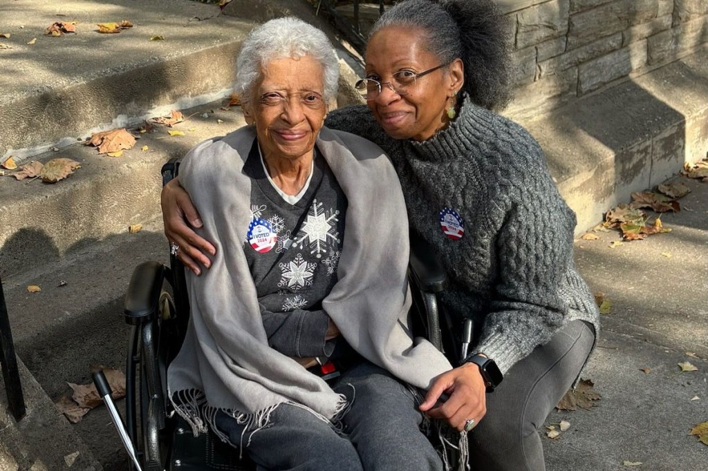 A portrait of two women side-by-side. On the viewer's left is a senior woman in a wheelchair. On the right is a middle-aged woman who has her arm around the senior woman as she kneels beside her. They are both smiling warmly.