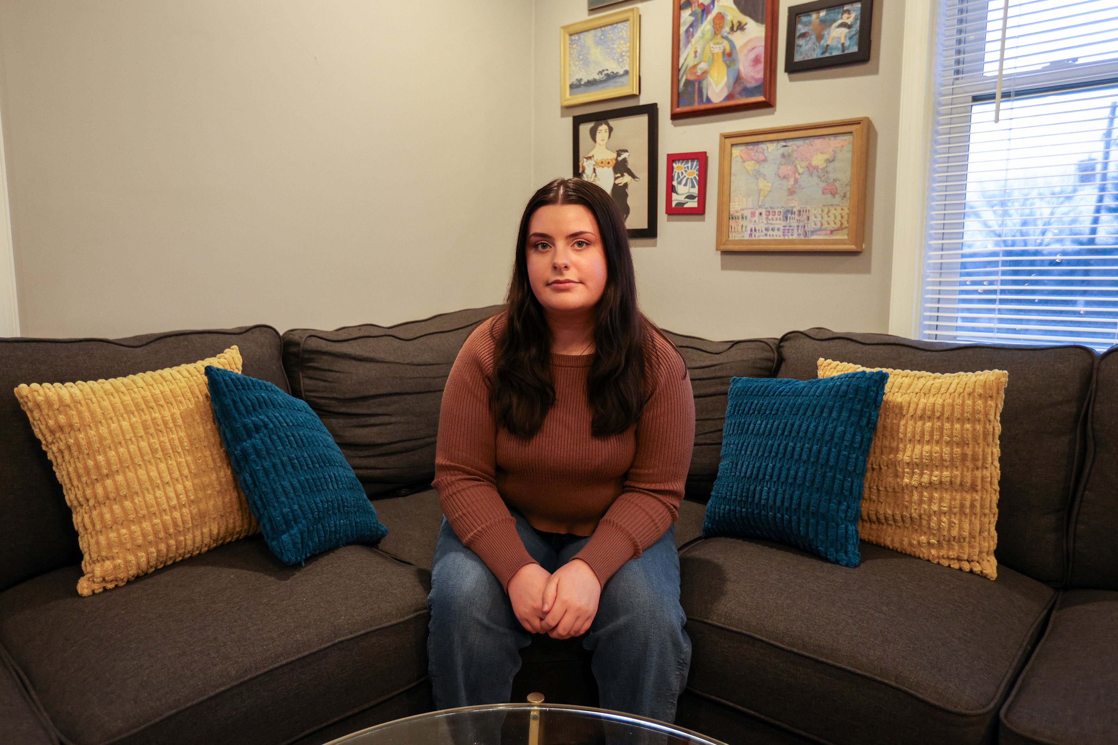 A 25-year-old woman sits on a couch and looks directly towards the camera.