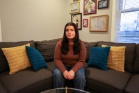 A 25-year-old woman sits on a couch and looks directly towards the camera.