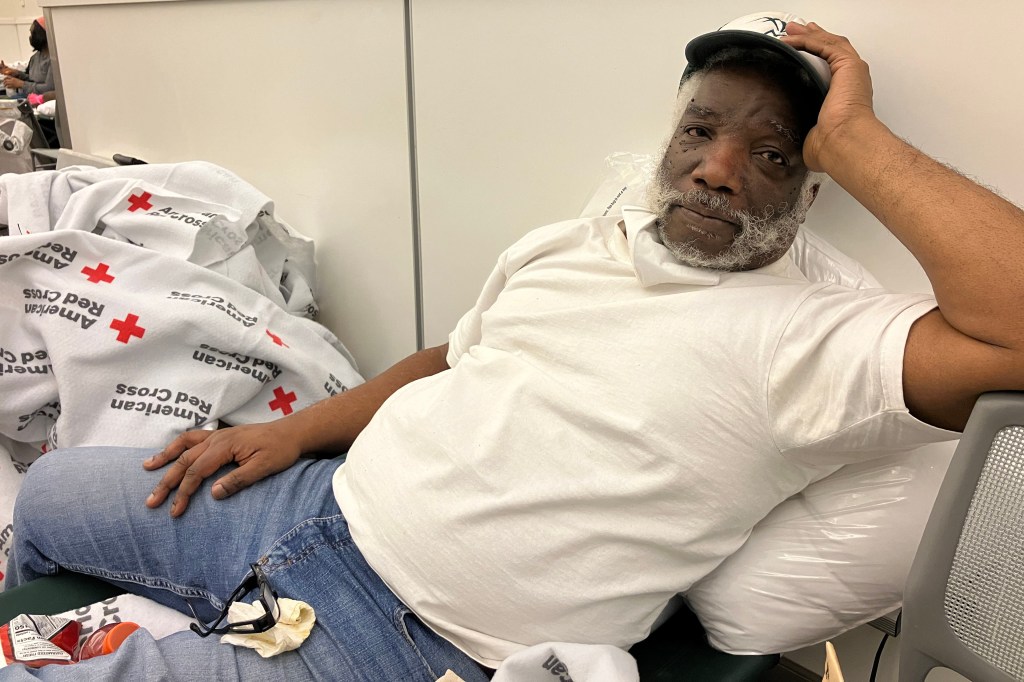 A man in a white shirt and jeans poses for a photo sitting in a chair next to an American Red Cross blanket