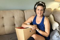 A woman with short brown hair held back with a white headband sits on a beige couch with a box in her lap filled with things. She is wearing long earrings and a blue tank top. Her legs are crossed and she is wearing knee-high black socks.