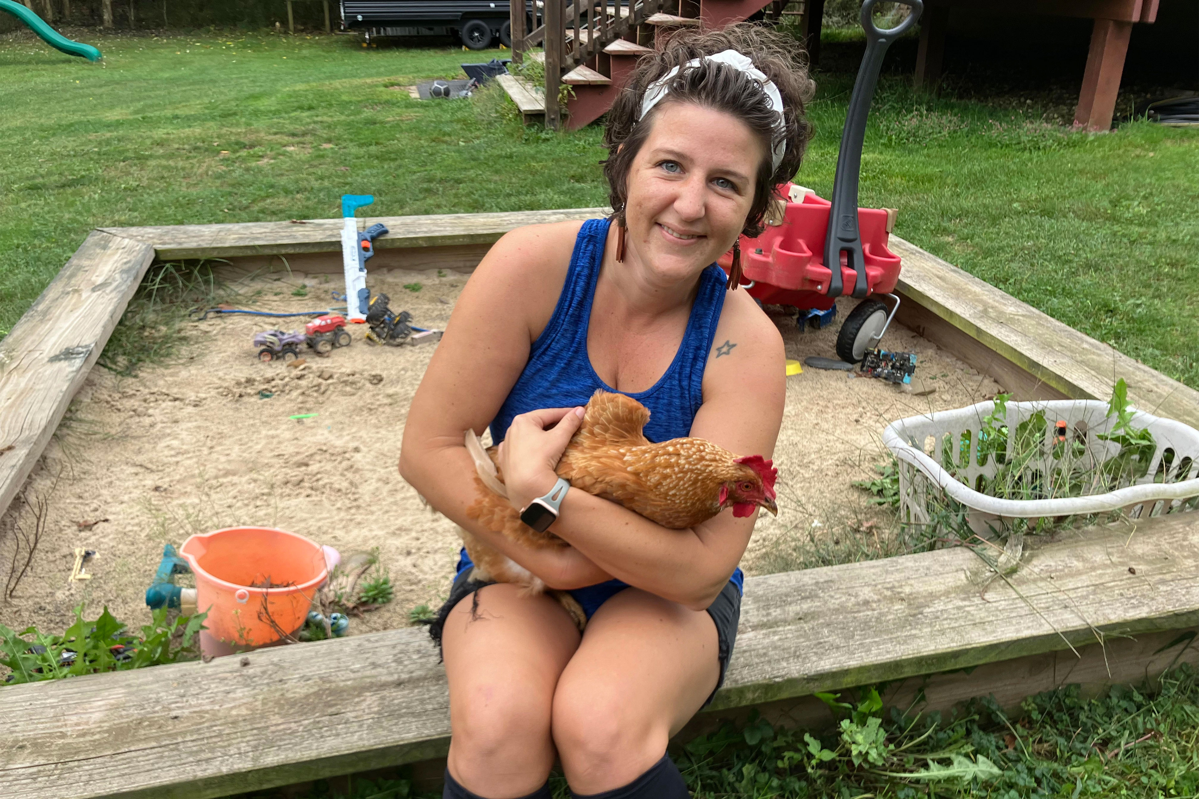 A woman with short brown hair held back with a white headband sits on the side of a wooden sand box cuddling a tan brown chicken. She is wearing long earrings and a blue tank top. Behind her, the yard is strewn with kids toys such as plastic buckets and trucks.