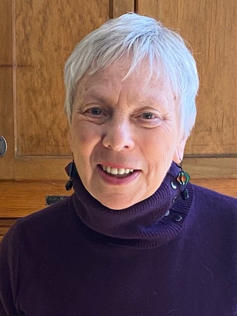 A senior woman with white short hair and a purple turtleneck sweater looks at the camera smiling