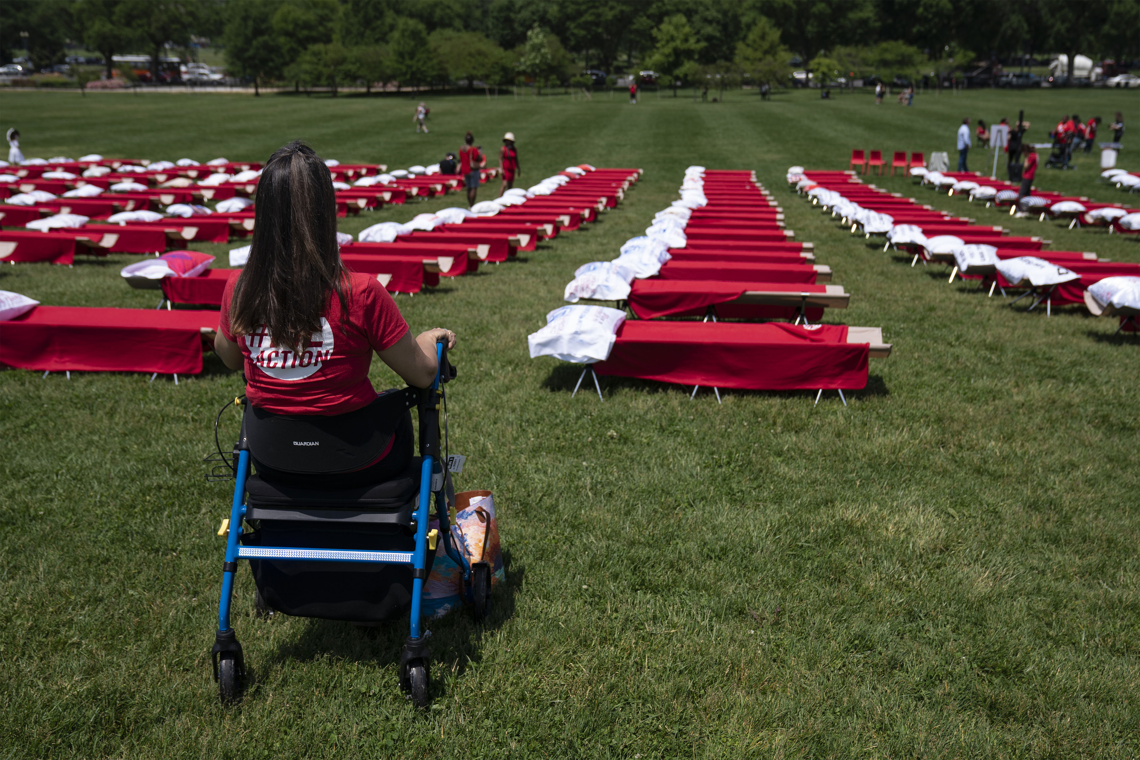 Across a green lawn, 500 cots with white pillows and red blankets are set up in rows. In the foreground, a woman in a red t-shirt with brown, long hair is seated on a blue and black walking aid device.