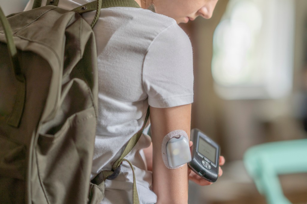 A young teen girl stands as she scans her diabetic patch on her arm with her phone. She is dressed casually and has a backpack on as she focuses on managing her diabetes.