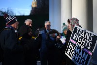 A small crowd of people face each other outdoors on a cold day. A man on the left wears a winter hat that says "TRUMP." A woman on the right holds a sign that says, "Trans rights are human rights!"