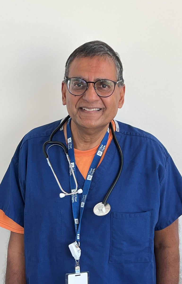 A portrait of Dr. Ravi Salgia. He wears blue scrubs and has a stethoscope hanging around his neck. He smiles at the camera.