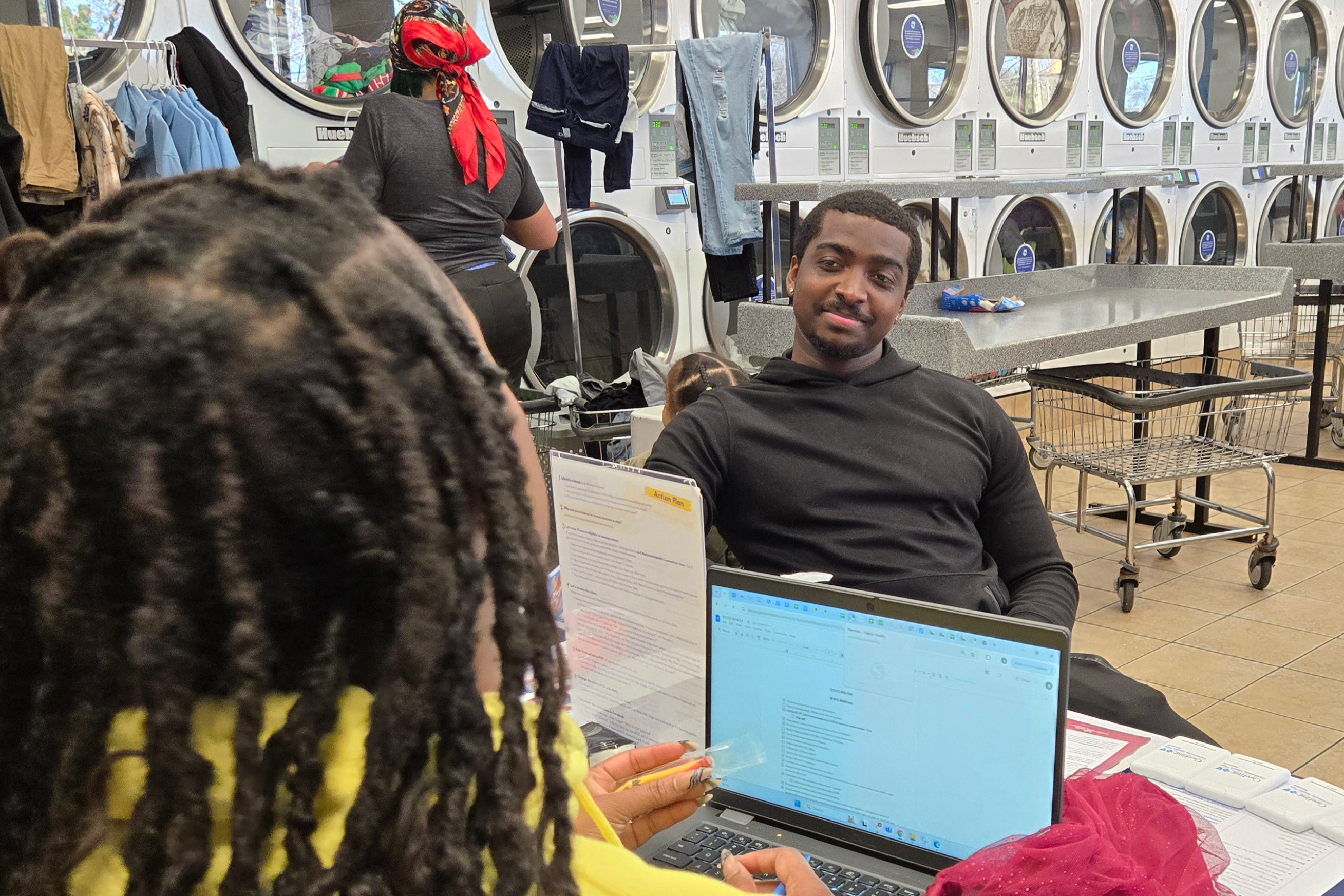 A photo of Brendan, Glover, a young man seated across from Adrienne Jones, who has a laptop in front of her. They are at a laundromat.