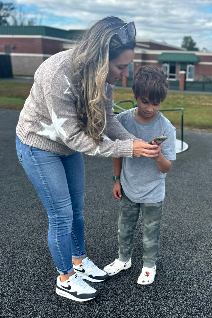 A mother stands beside her elementary-aged son. They are looking at a cell phone that has an app that is connected to the child's continuous glucose monitor.