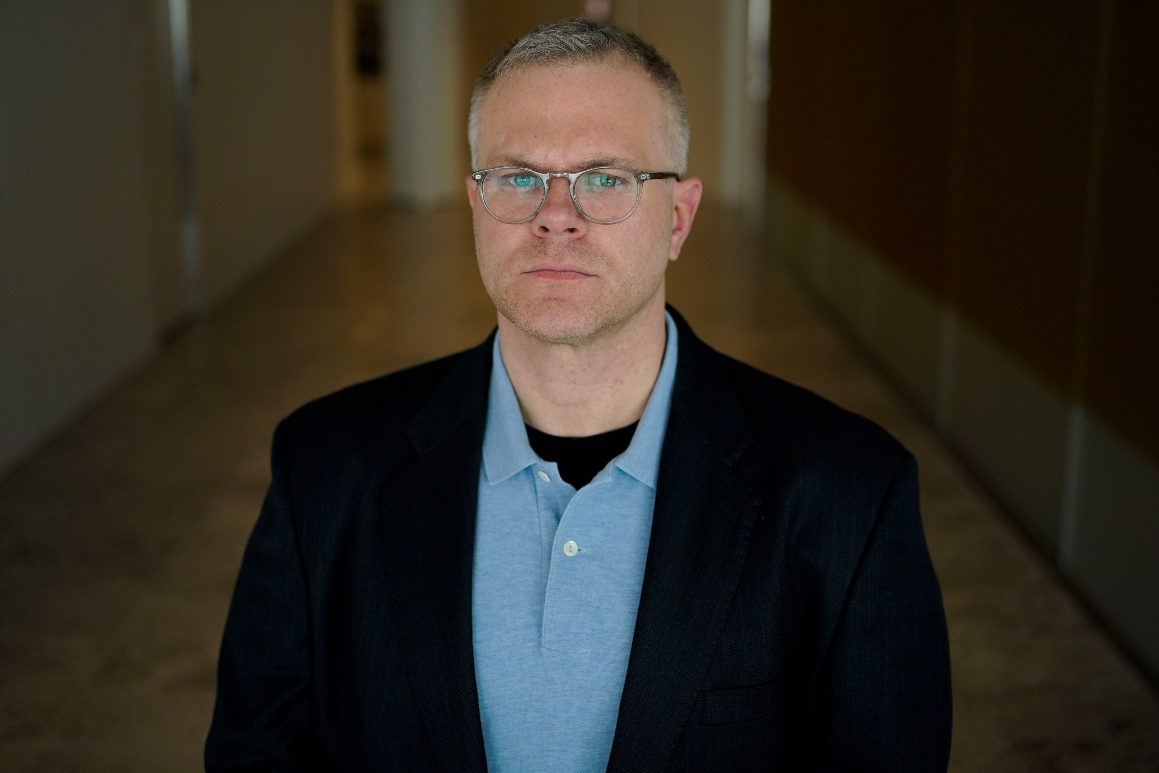 A photo of a man in glasses standing for a portrait in a hallway.