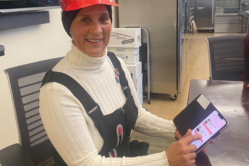 A woman wearing overalls and an orange hard hat is sitting for a photo. She holds a cell phone in her hand for the camera to see and smiles in the direction of the photographer.