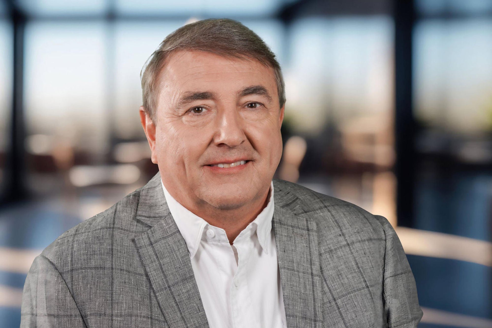 A man in a white collared shirt and gray patterned blazer smiles at the camera. The room behind him is in such soft focus that you can't make out any details.