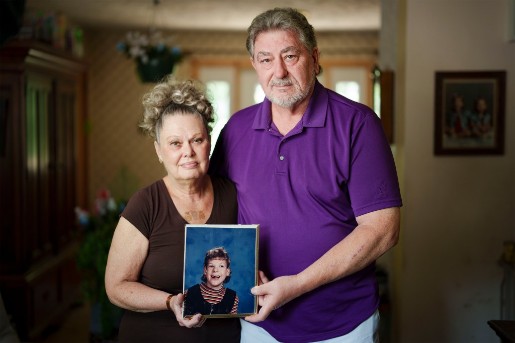 A photo of an older man and older woman standing for a photo in their living room. They're holding a school photo of their daughter.