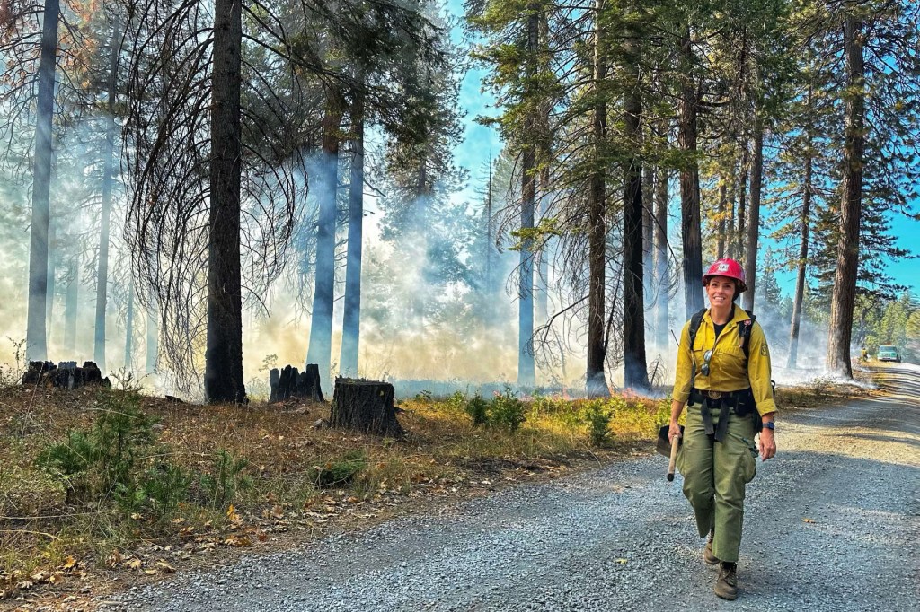 A female firefighter is walking on a road with a forest, hazy with smoke highlighted by afternoon sun, behind her.