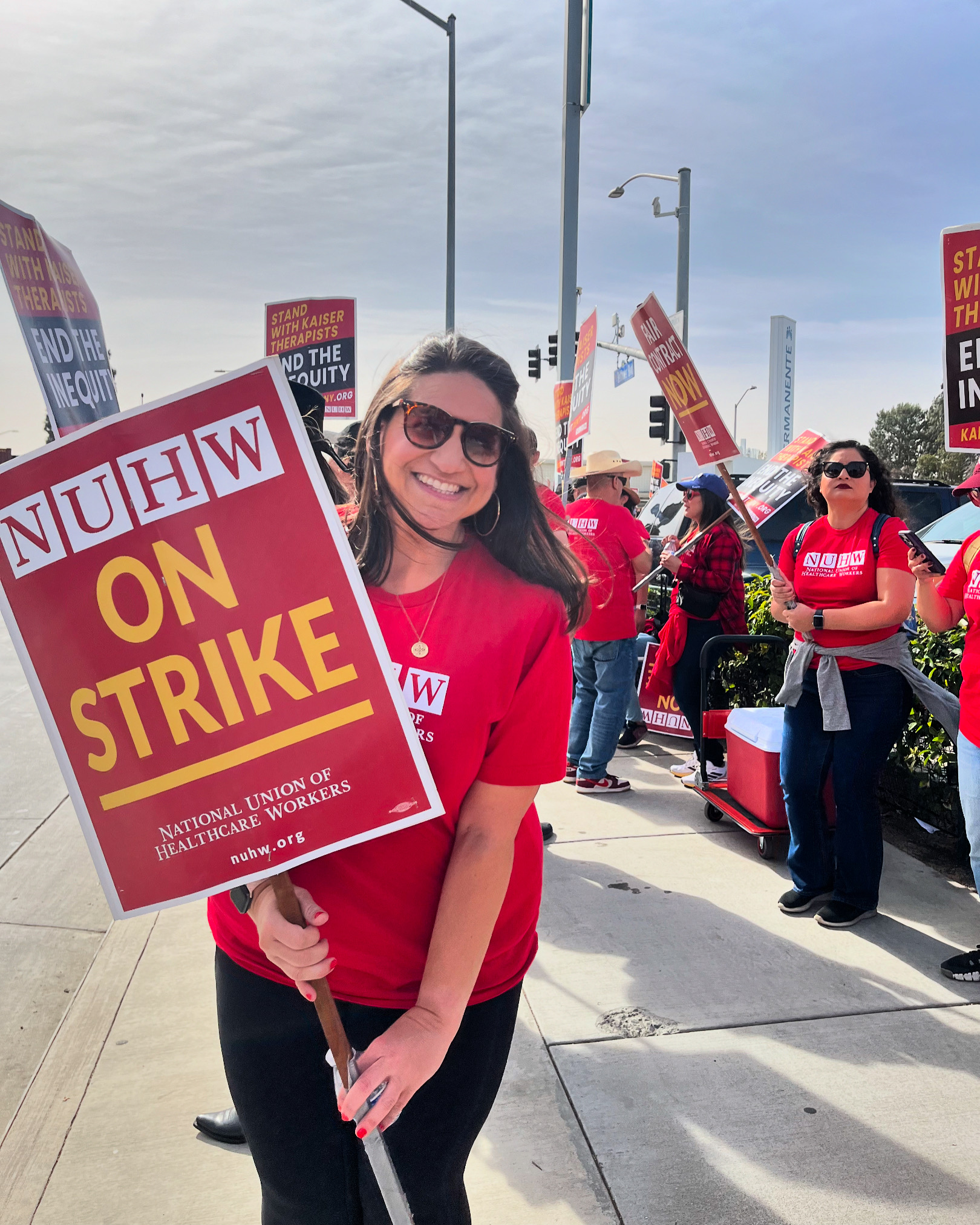 A photo of Kassaundra Gutierrez-Thompson posing for a photo with her fellow workers on strike. She holds a sign that reads, "NUHW On Strike."