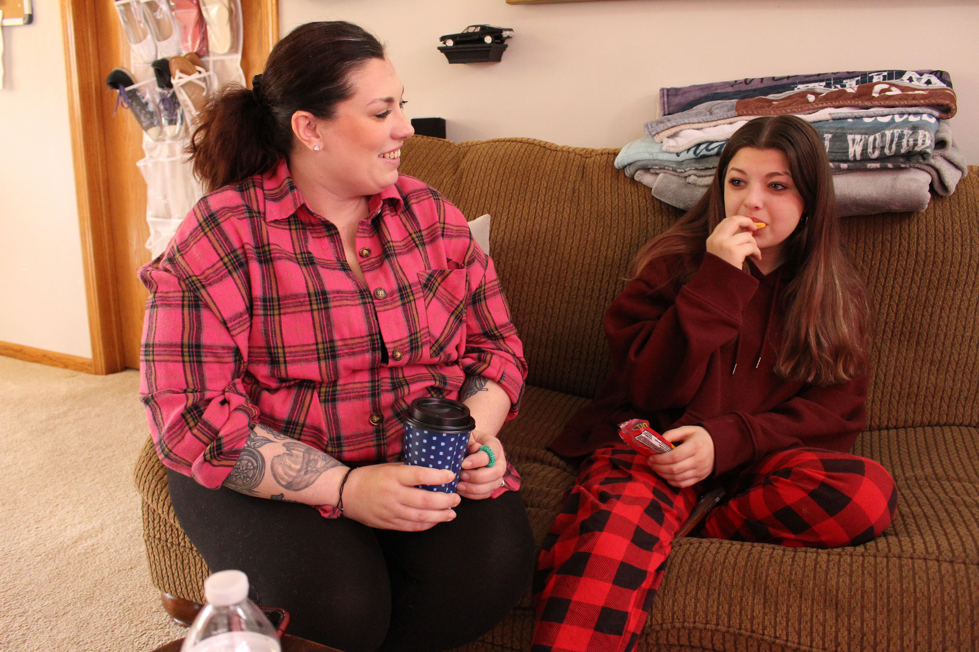 A woman in a pink plaid shirt holds a coffee cup and sits on a couch next to a girl in a burgundy hoodie sweatshirt