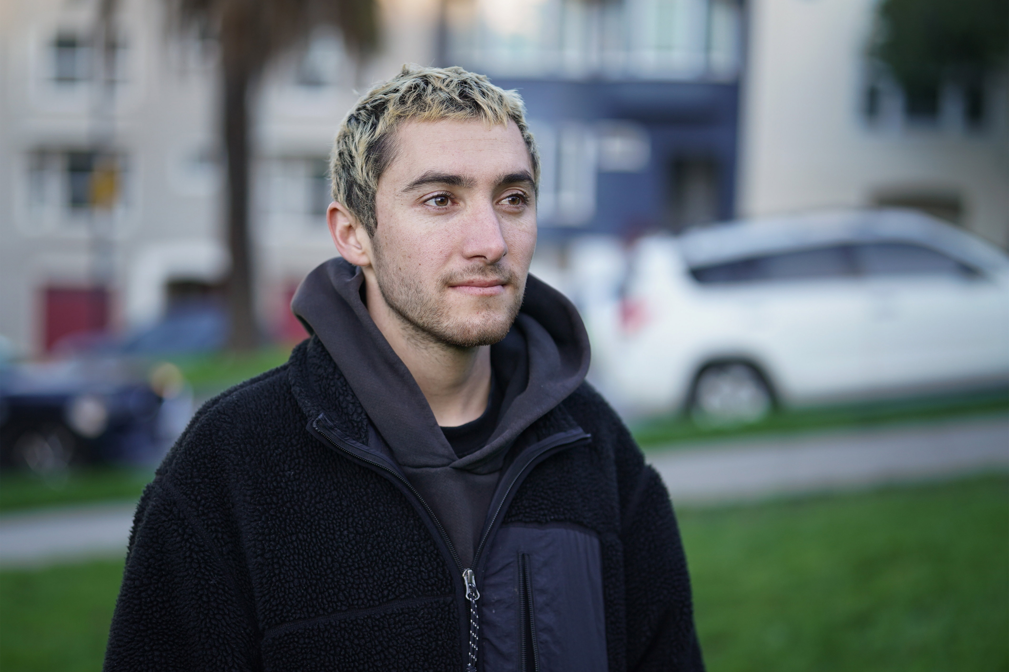 A photo of a young man posing for a portrait outside. A white car drives up the street behind him.