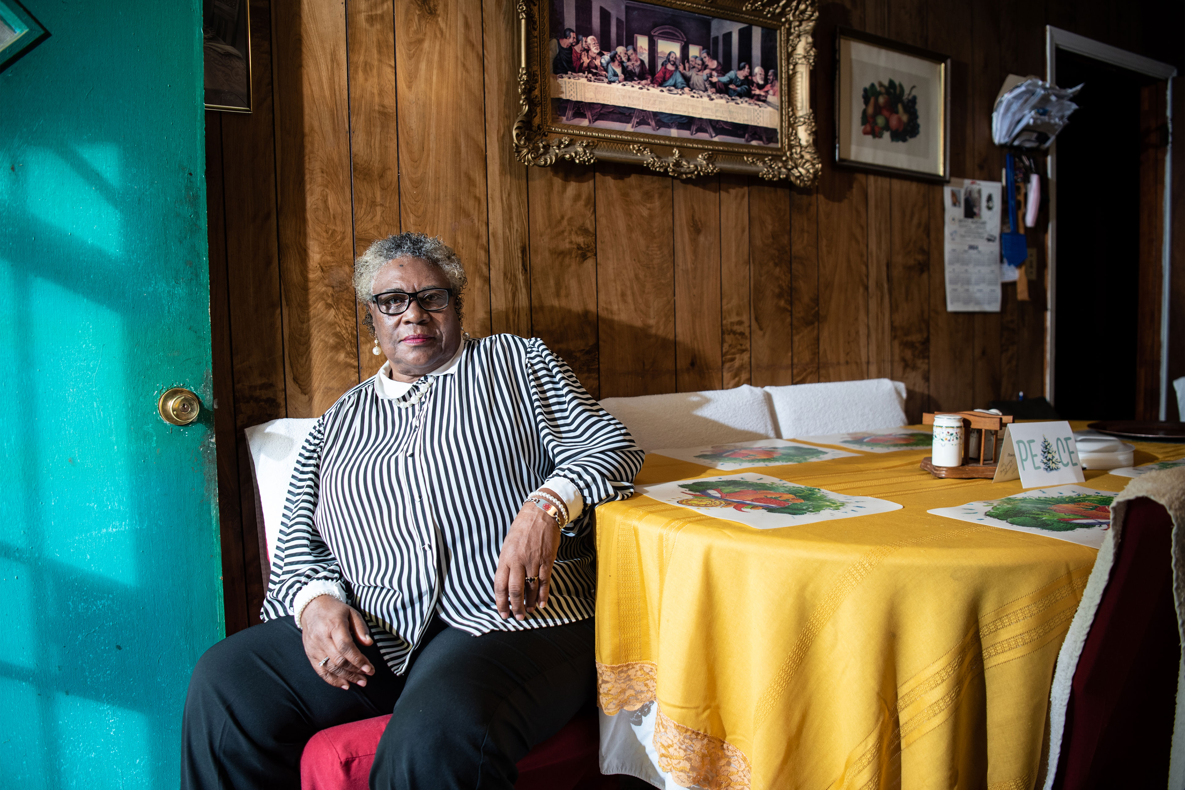 A woman with short curtly hair and glasses looks at the camera and sits beside a table covered in a yellow tablecloth. She wears a blank and white striped blouse and rests her left elbow on the table. A teal door is open just to her right.