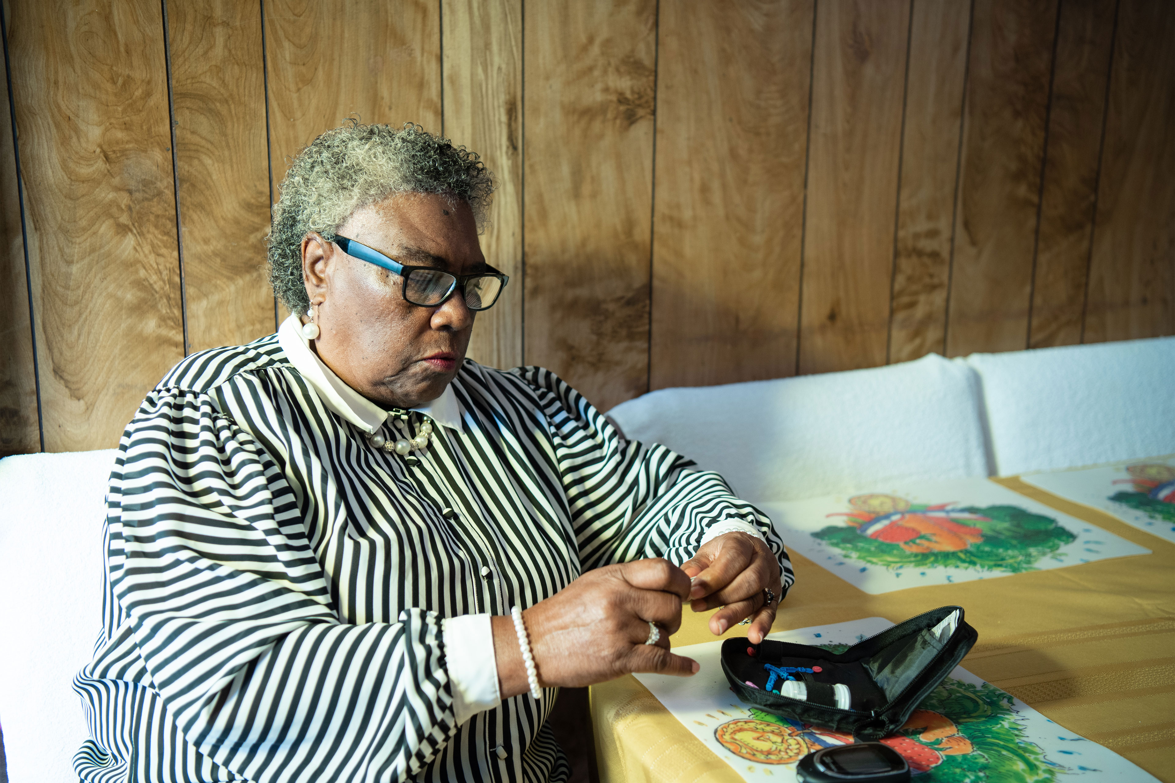 An image of a woman sitting at a table preparing to test her blood glucose. She has short curtly hair and glasses, and wears a black and white striped blouse. A zippered pouch sits on the table beside her.