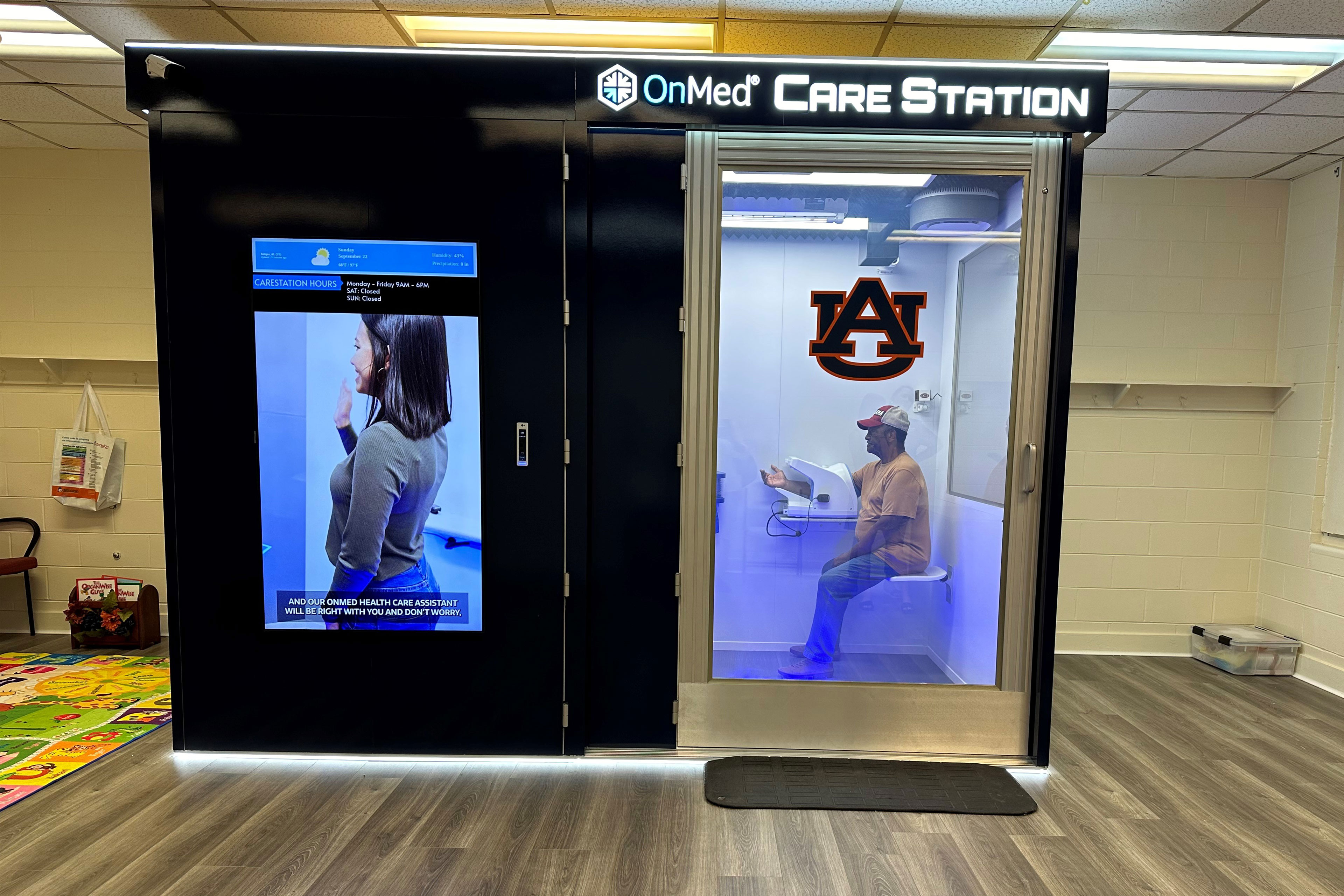 A booth with a large screen on the left and a door with a window on the right and the text "OnMed Care Station" at the top. Through the window can be seen a man in a t-shirt, jeans, and cap sitting inside a booth with his right arm in a device.