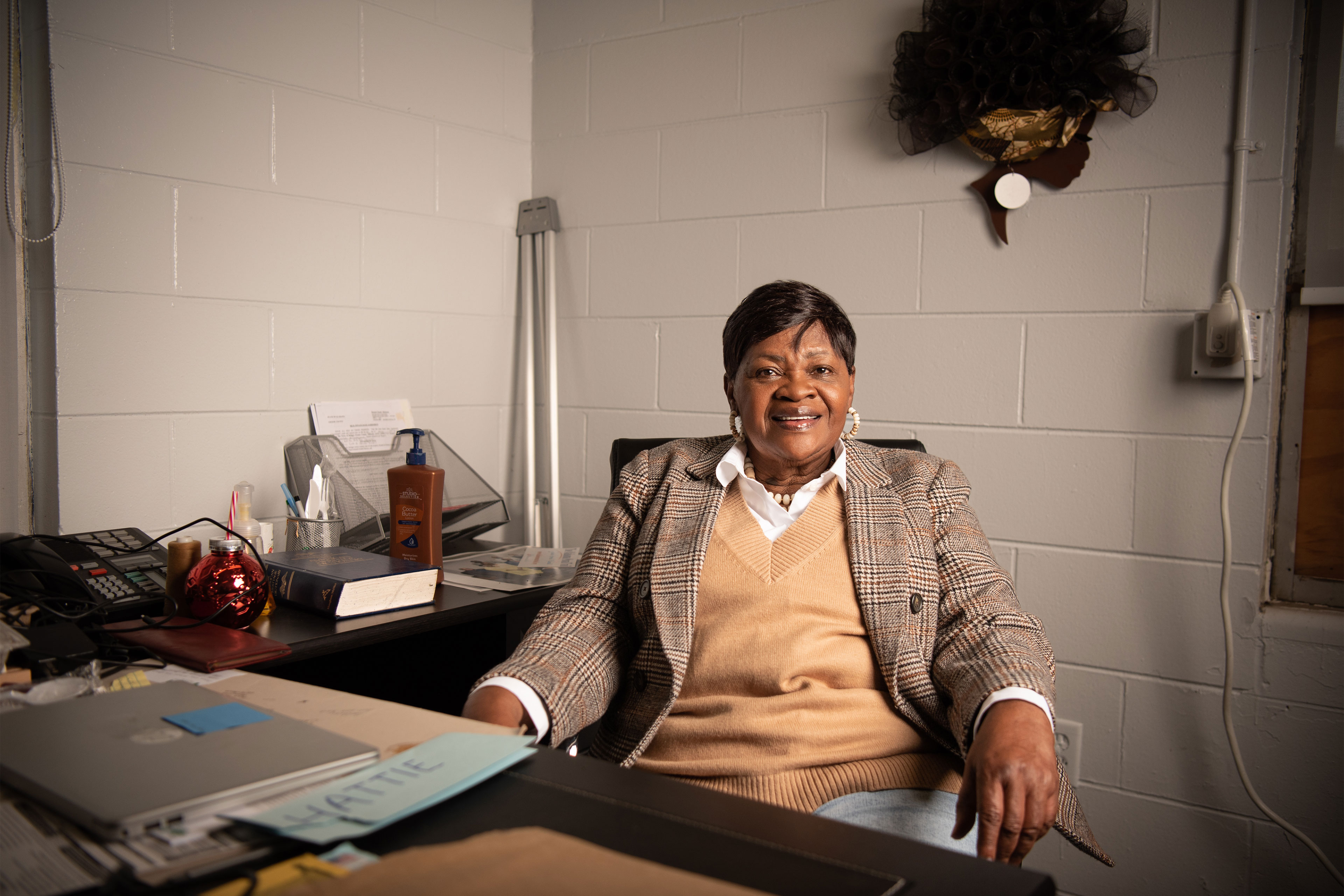 A woman wearing a plaid blazer over a collared shirt and sweater sits behind a desk and smiles at the camera.