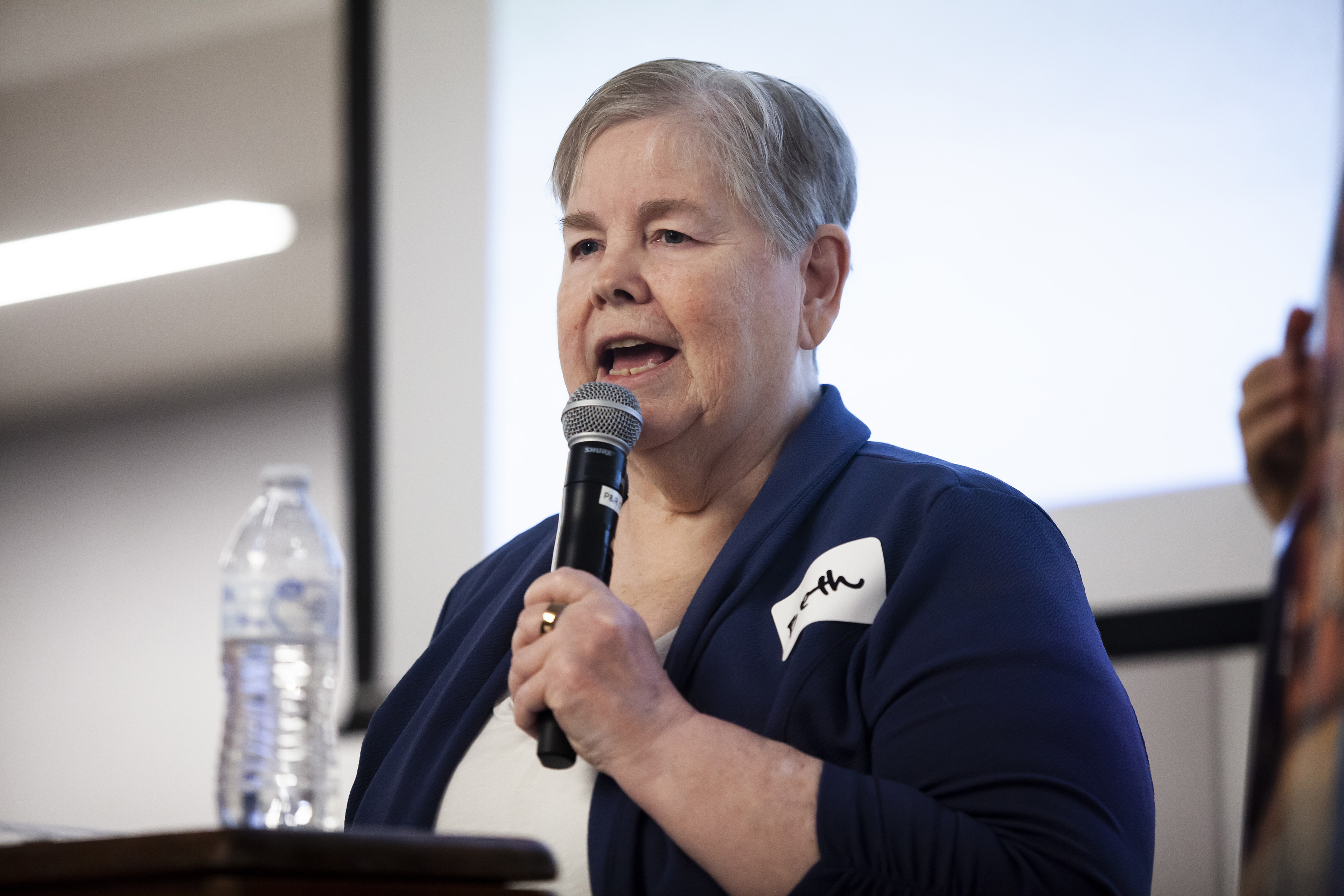 A woman with short, gray hair, wearing a navy sweater, speaks at a podium.