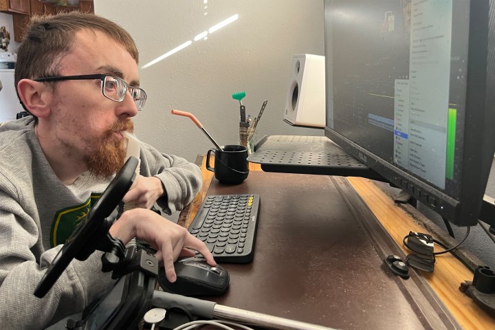 A photo of a man seated at a desk, working on a computer, breathing through a portable ventilator.