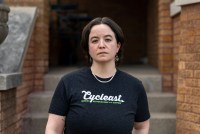 An adult with short brown hair, wearing a black T-shirt, stands in front of a stoop and is directly facing the camera.