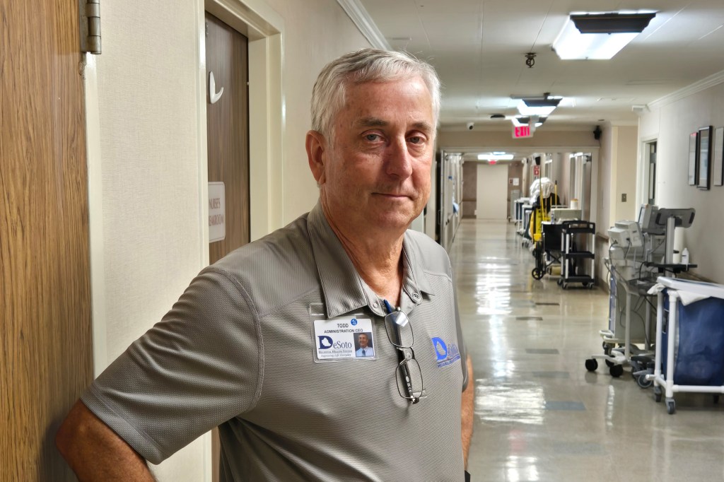A photo of an older man standing for a portrait in a hospital corridor.