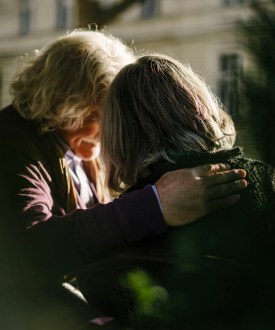 An unidentifiable grey haired senior couple are photographed from behind, their faces obscured from view. One has their hand on the other's shoulder.