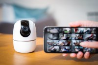 A photo of a small video camera device resting on a table. A hand holds up a phone screen in front of it, showing that camera feed can be monitored on the phone.