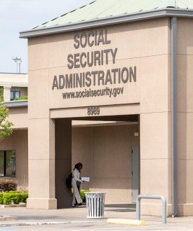 A photo of a concrete office building with the words "Social Security Administration" and "www.socialsecurity.gov" on a sign on the front entrance. An American flag on a pole is posted near the entrance.
