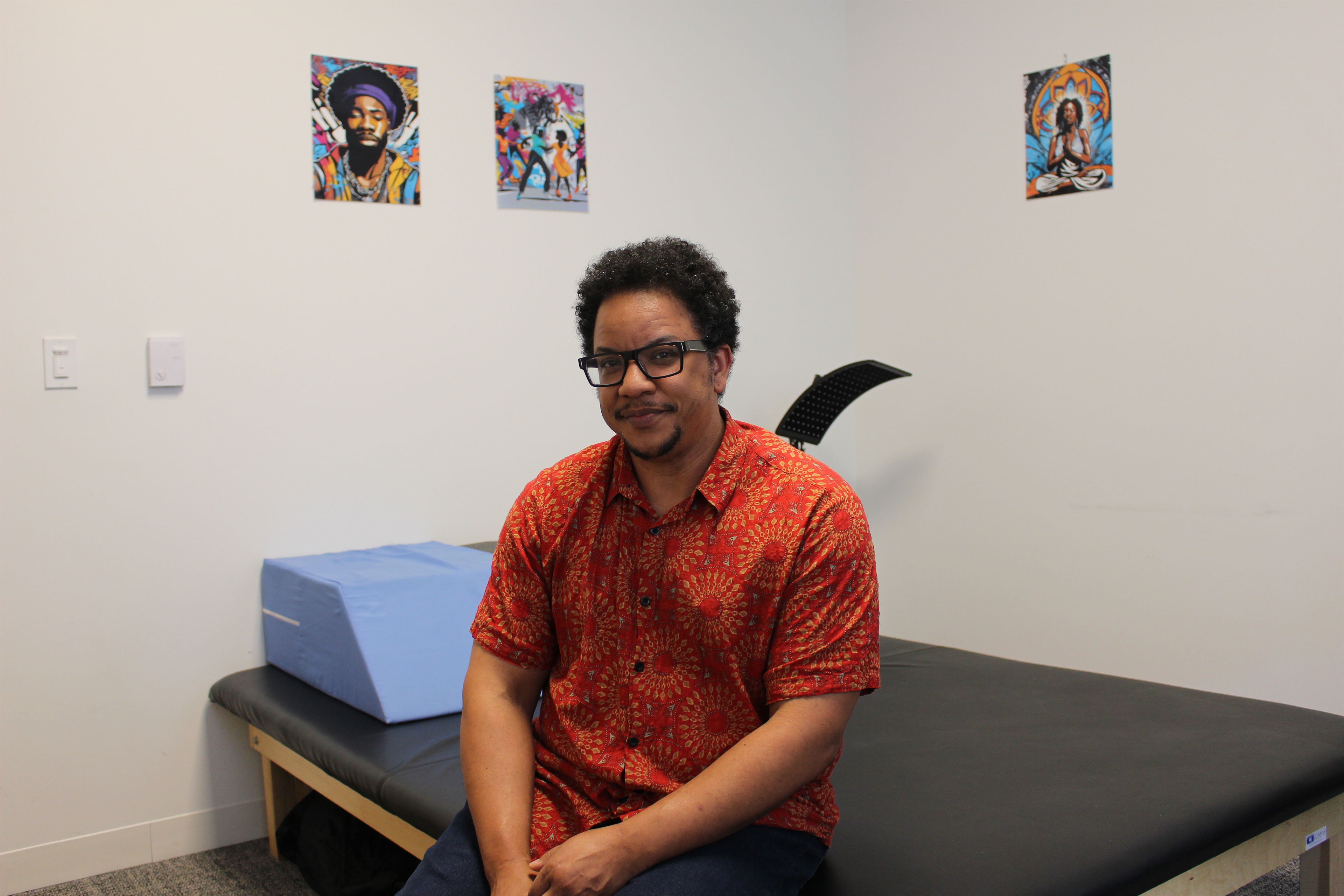 A man wearing dark glasses, an orange and red print collared short-sleeve shirt, and dark pants sits on a low padded table and looks at the camera.