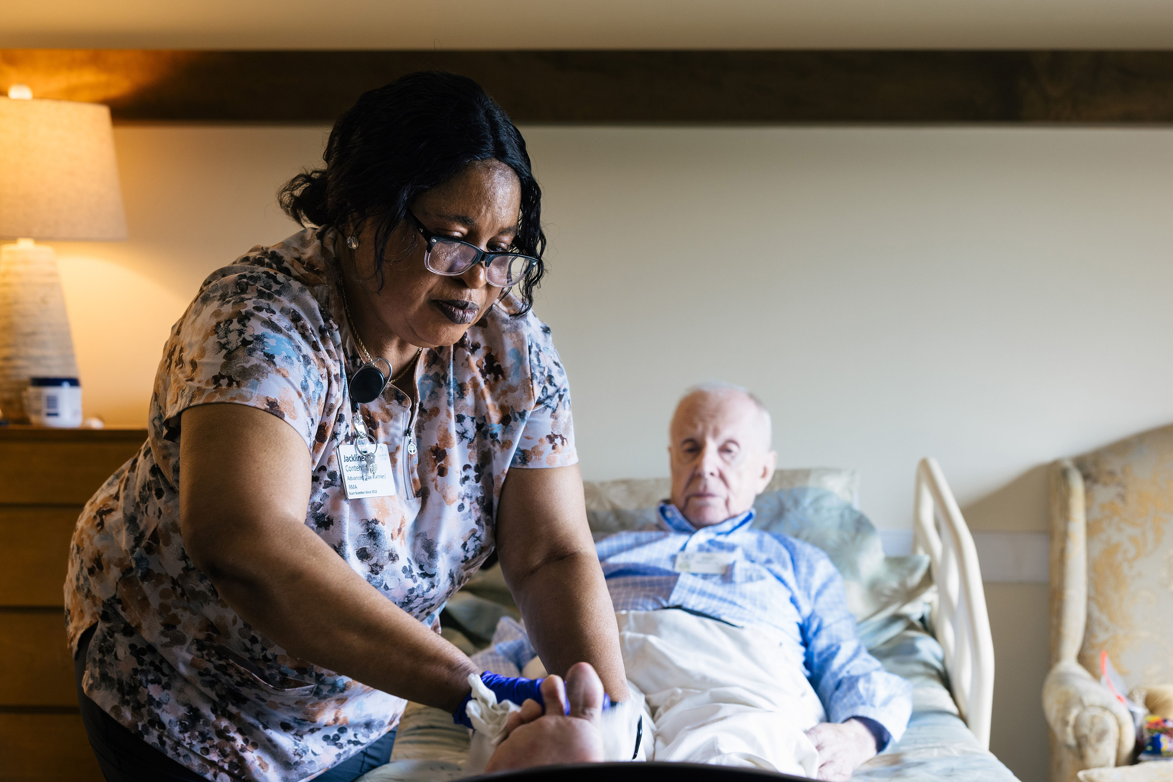 A senior man is lying in a bed in a nursing home. A nurse is carefully helping him with his socks.