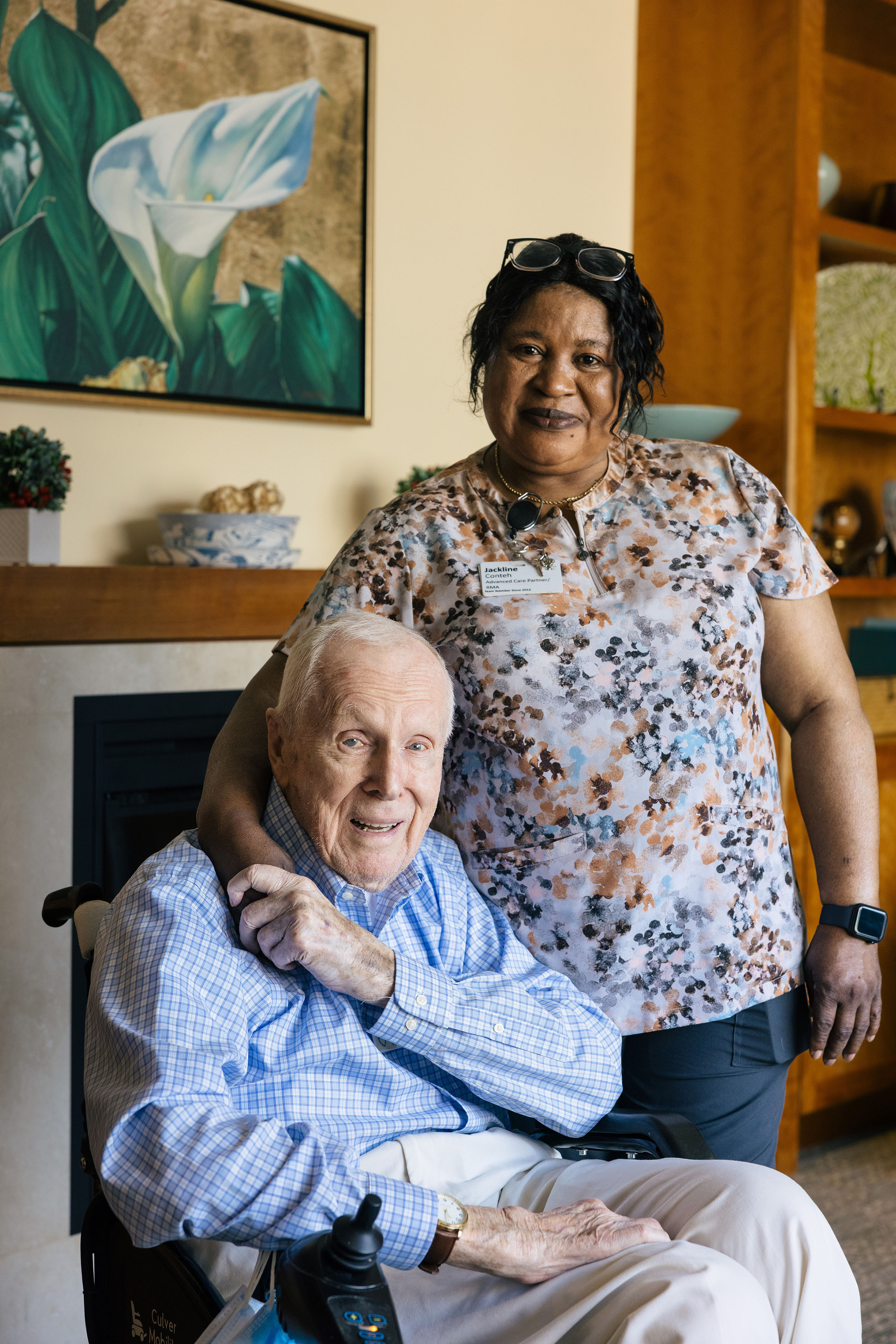 A senior man sits in a wheelchair, smiling wide as he grips the hand of his nurse, which is resting on his shoulder. The nurse stands beside him, also smiling warmly.