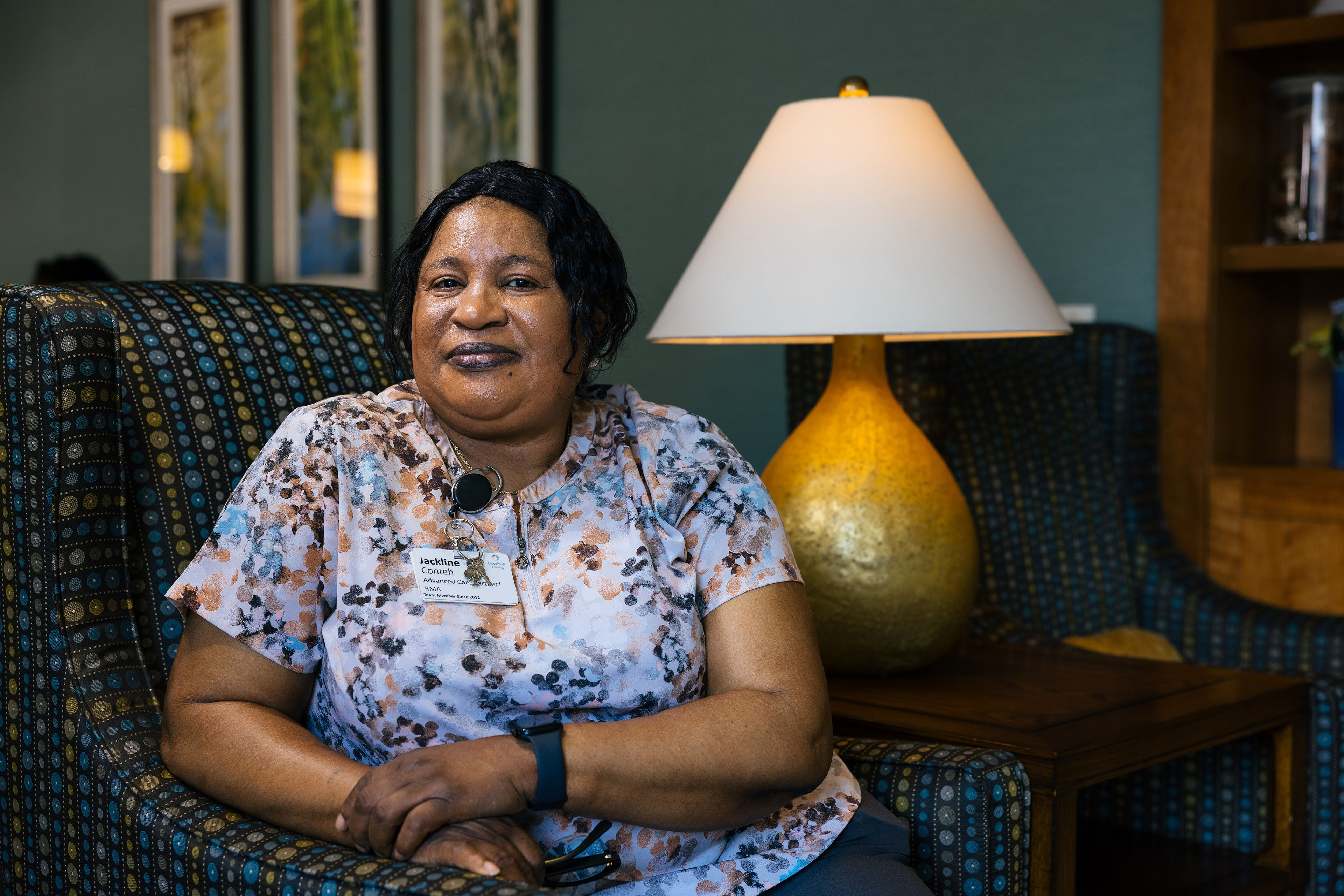 A woman wearing a floral shirt and a name-tag sits in a chair for a portrait.
