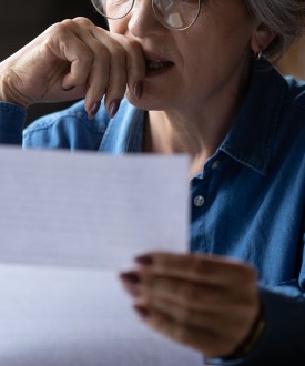 A closely cropped photo of a senior woman holding a paper letter. She presses her hand to her lips as she makes a stressed expression.