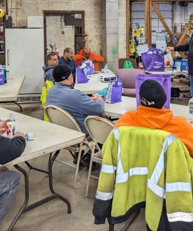 A man stands before a room of seated construction workers wearing high-vis clothing and hard hats.