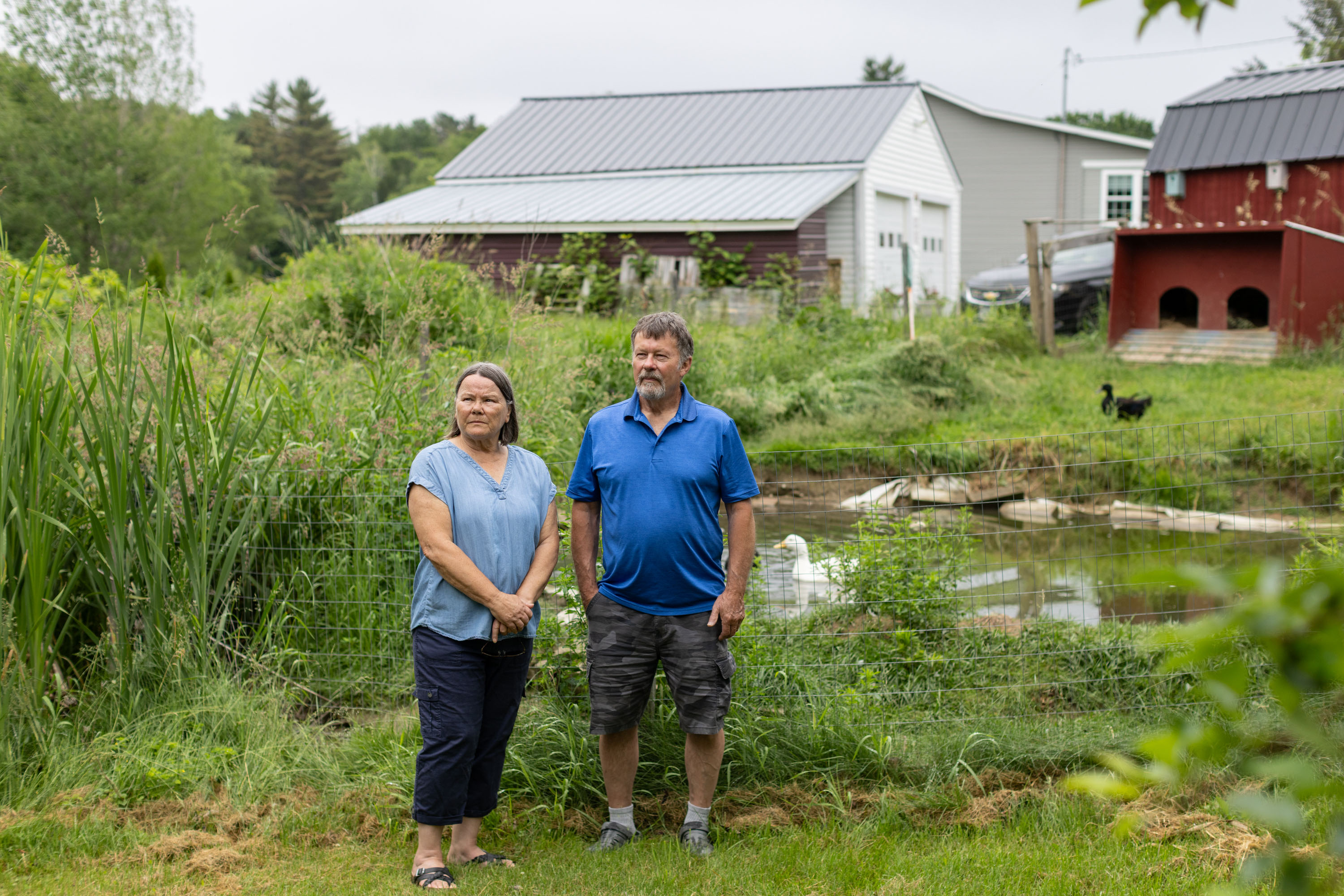 A husband and wife pose for a photo by their duck pond. The area is lush with green grasses and various plants.